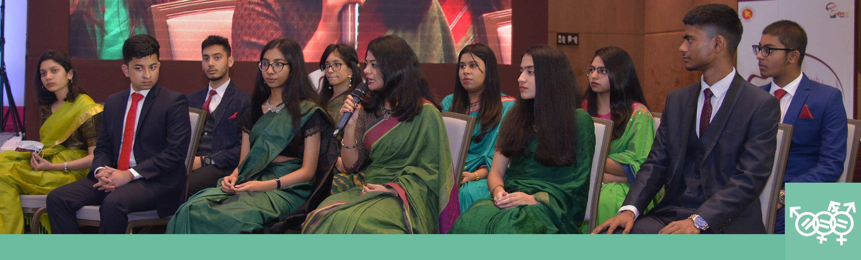 a group of young adults seated in two rows of chairs, one speaking from a microphone